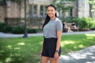 A young woman with a Warrior-Scholar Project shirt poses in front of a building on campus.