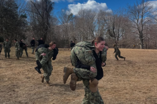 Two women in Navy working uniforms buddy-carry simulated casualties.